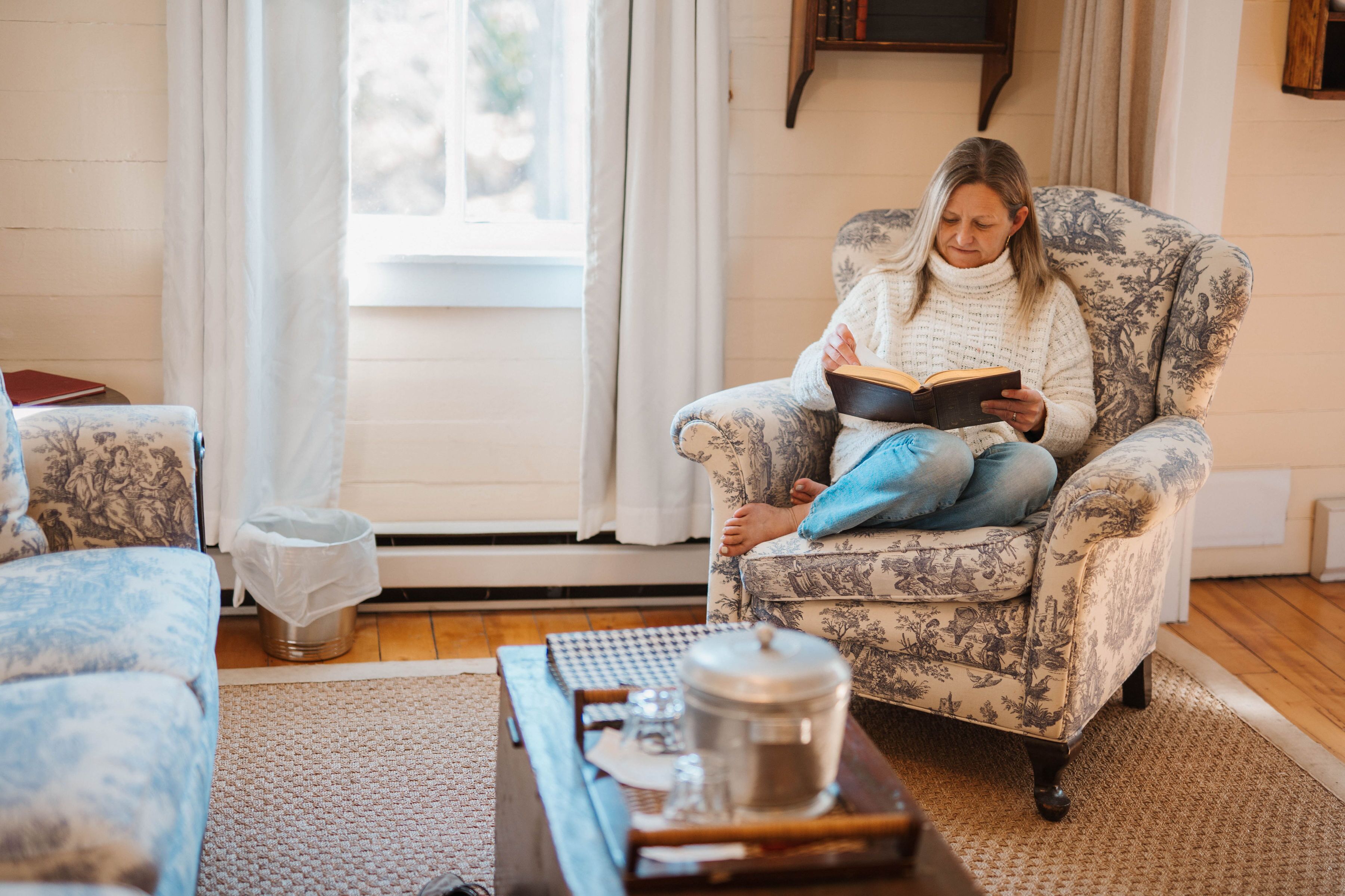 A person sitting on a floral-patterned armchair reading a book in a room with a wooden floor, a window with curtains, and a small table with a jar and a book.