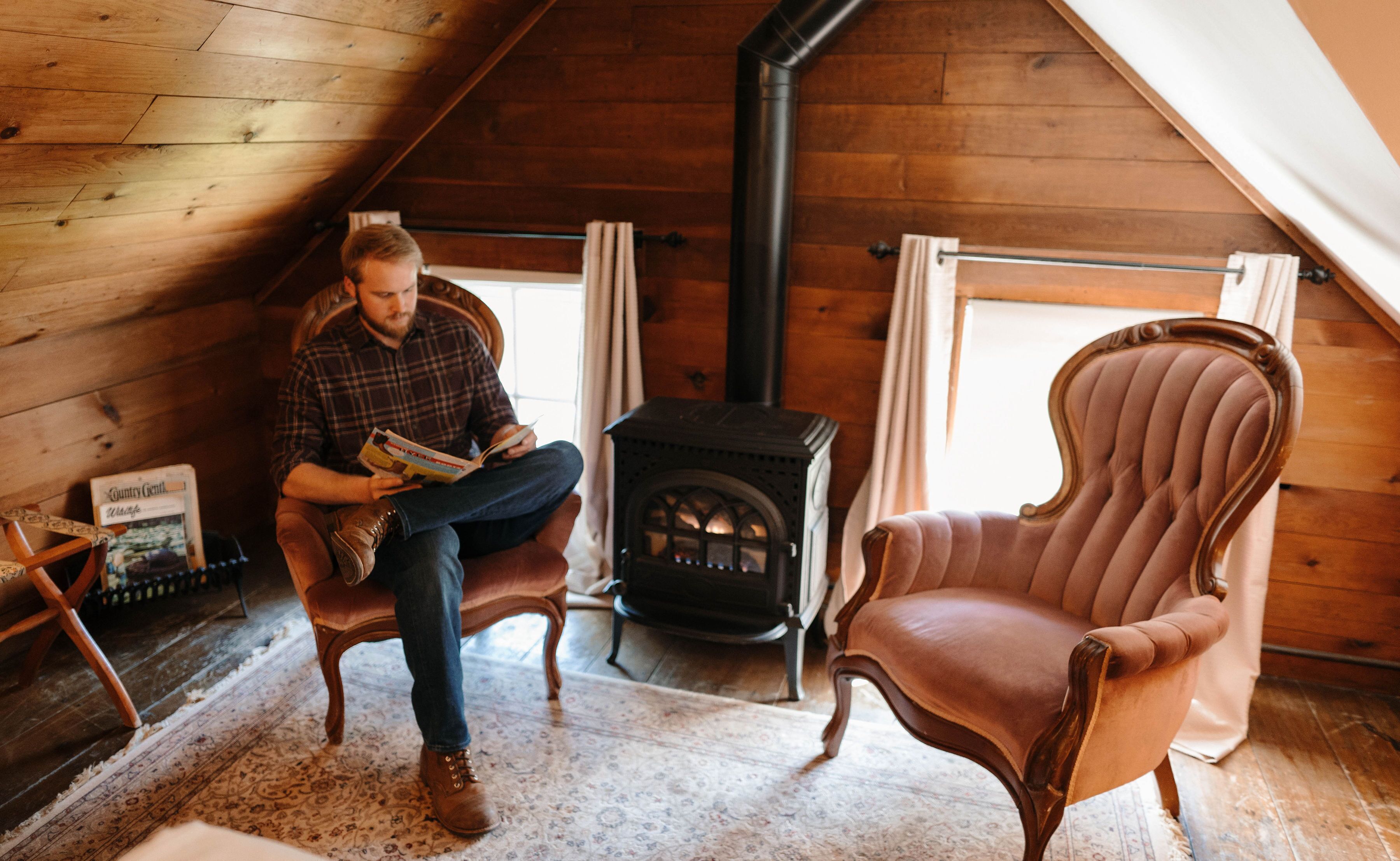 Una habitación acogedora con techo de madera, chimenea, sillón de cuero y una persona leyendo un libro.