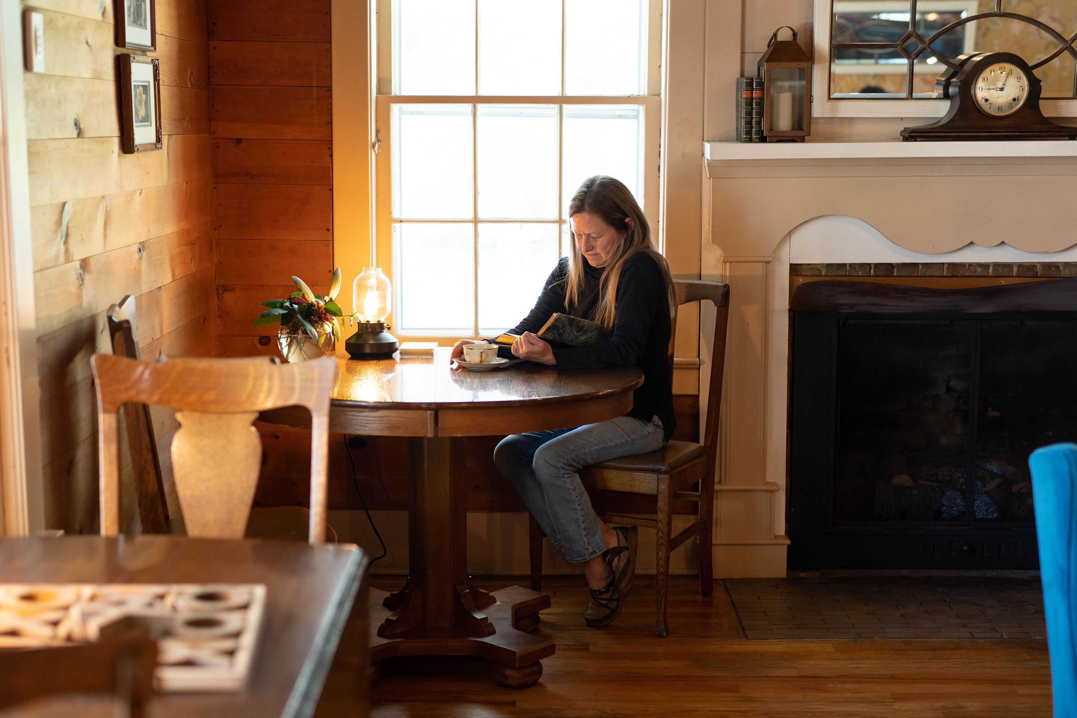 Une femme est assise à une table en bois près d’une cheminée et lit un livre.