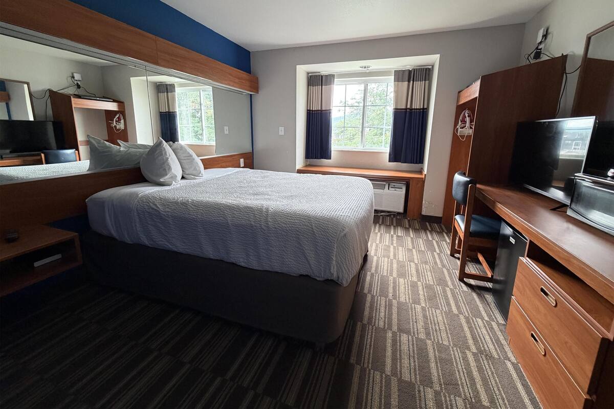 A hotel room with a bed, a desk with a television, a window with curtains, and a mirror.