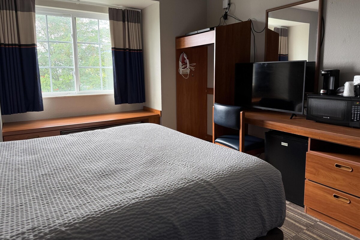 A hotel room with a bed, a desk with a television and microwave, a chair, a wardrobe, and a window with curtains.