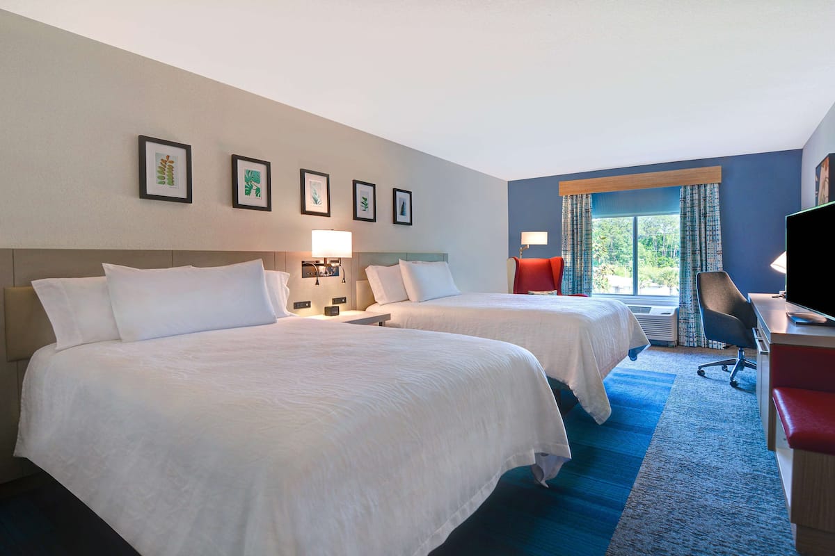 A hotel room with two beds, a desk, a TV, and a window with curtains.