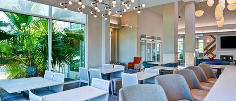 A modern indoor space with tables and chairs, large windows, and indoor plants.
