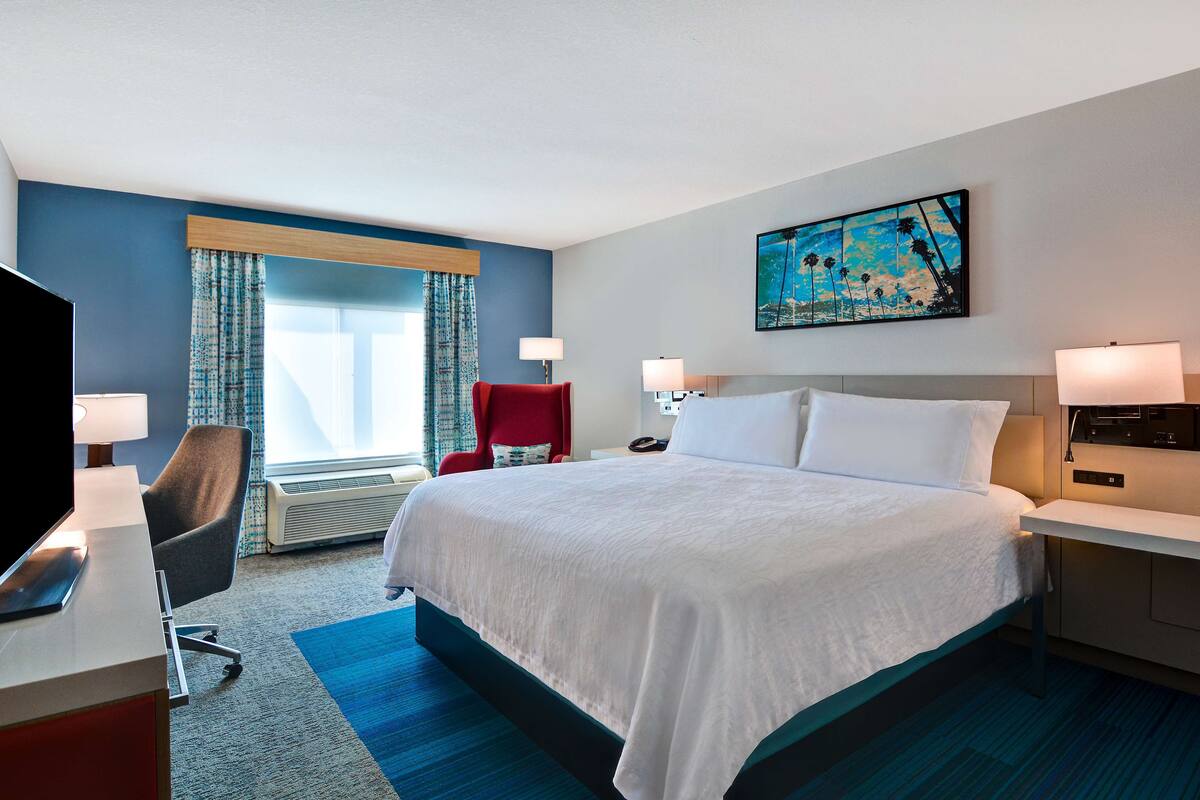 A hotel room with a large bed, a desk with a computer, a red chair, and a painting on the wall.