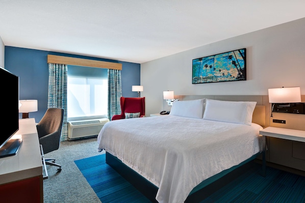 A hotel room with a large bed, a desk with a computer, a red chair, and a painting on the wall.