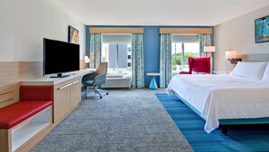 Habitación de hotel con cama, escritorio con televisor, una silla roja y ventana con cortinas.