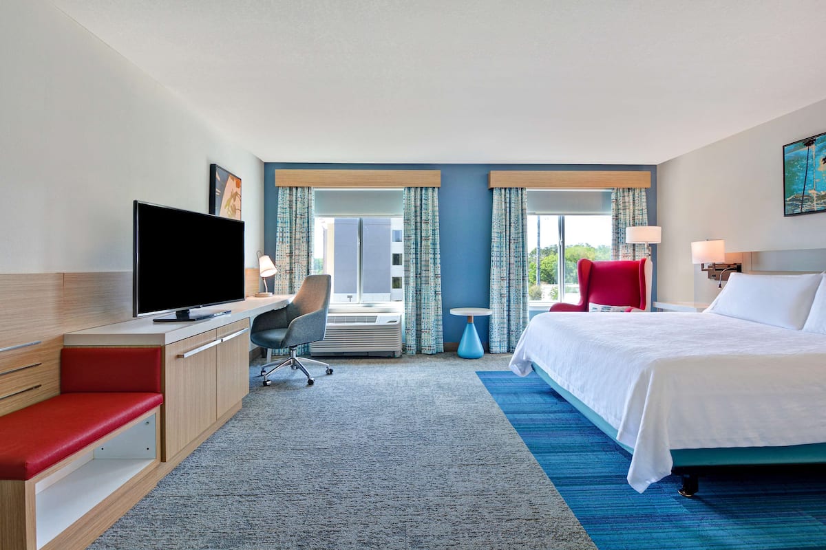 A hotel room with a bed, a desk with a TV, a red chair, and a window with curtains.