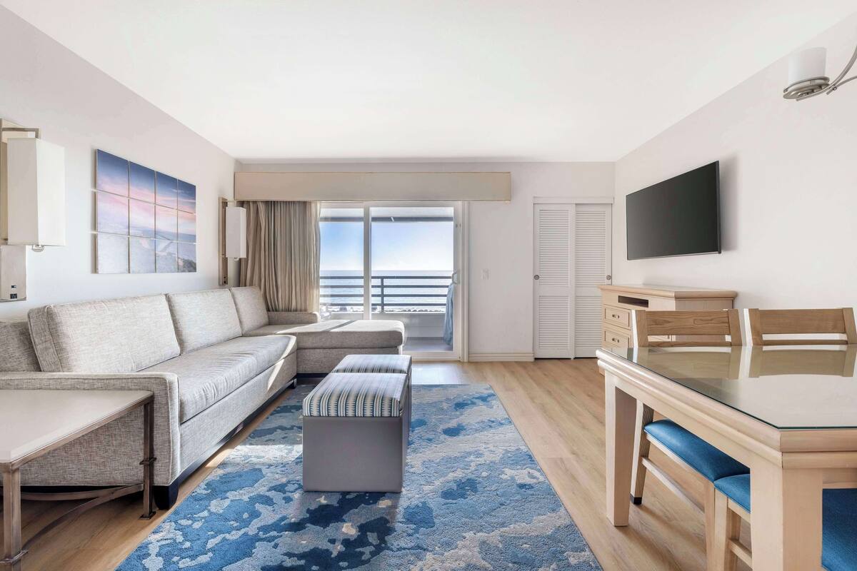 Ein modernes Hotelzimmer mit einer Couch, Hocker und einem Esstisch mit Stühlen. Ein großes Fenster bietet einen Blick ins Freie.