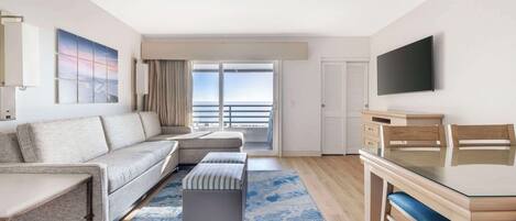 Una habitación de hotel moderna con sofá, reposapiés y una mesa de comedor con sillas. Una ventana grande ofrece vistas al exterior.