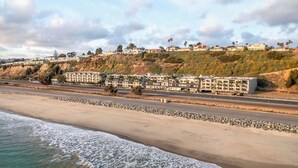 Un hotel frente al mar con varias construcciones, una playa en primer plano y un paisaje montañoso al fondo.