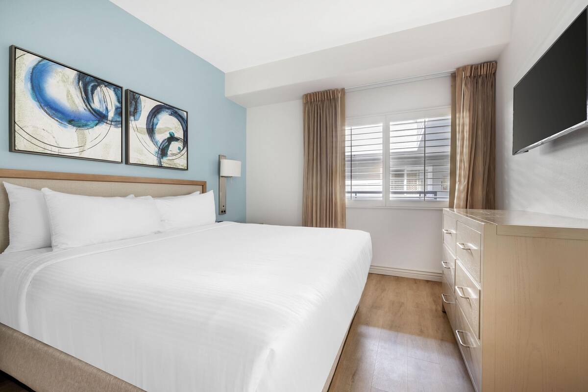 Ein Hotelzimmer mit einem großen Bett, einem Flachbildfernseher, einem Schminktisch und abstrakten Wandbildern.