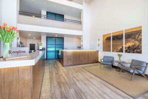 A hotel reception area with a wooden reception desk, a glass door leading to a balcony, and a seating area with two chairs and a small table. - Hilton Vacation Club Riviera Beach & Shores (Dana Point)