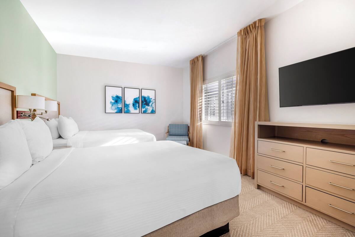 Ein Hotelzimmer mit einem großen Bett, einem Schreibtisch, einem Sessel, einem Fernseher und Kunstwerken an der Wand.
