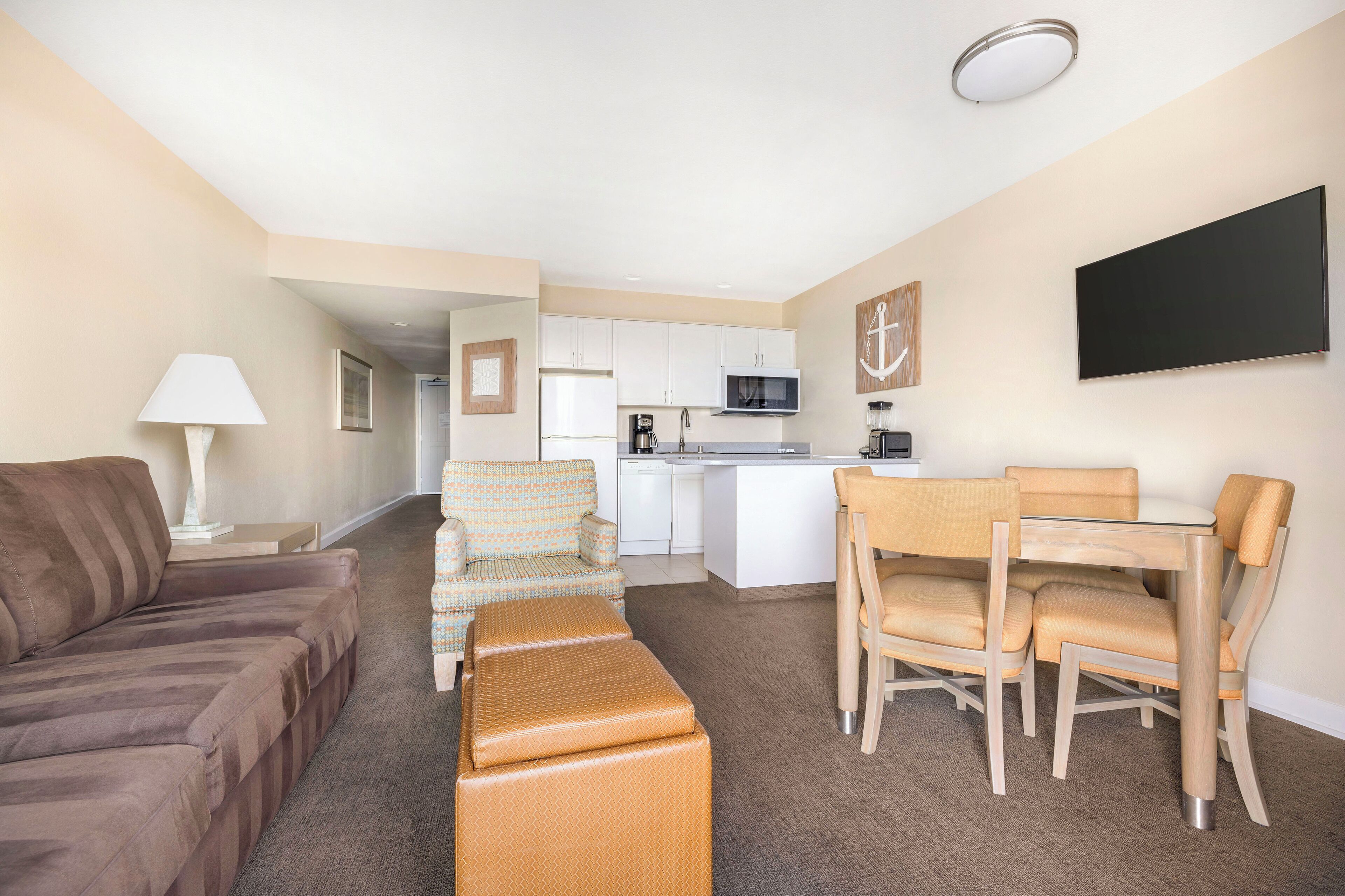A compact living space with a sofa, a patterned armchair, a dining table with chairs, a kitchenette, and a flat-screen TV.