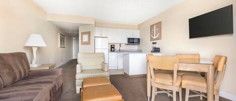 A compact living space with a sofa, a patterned armchair, a dining table with chairs, a kitchenette, and a flat-screen TV.