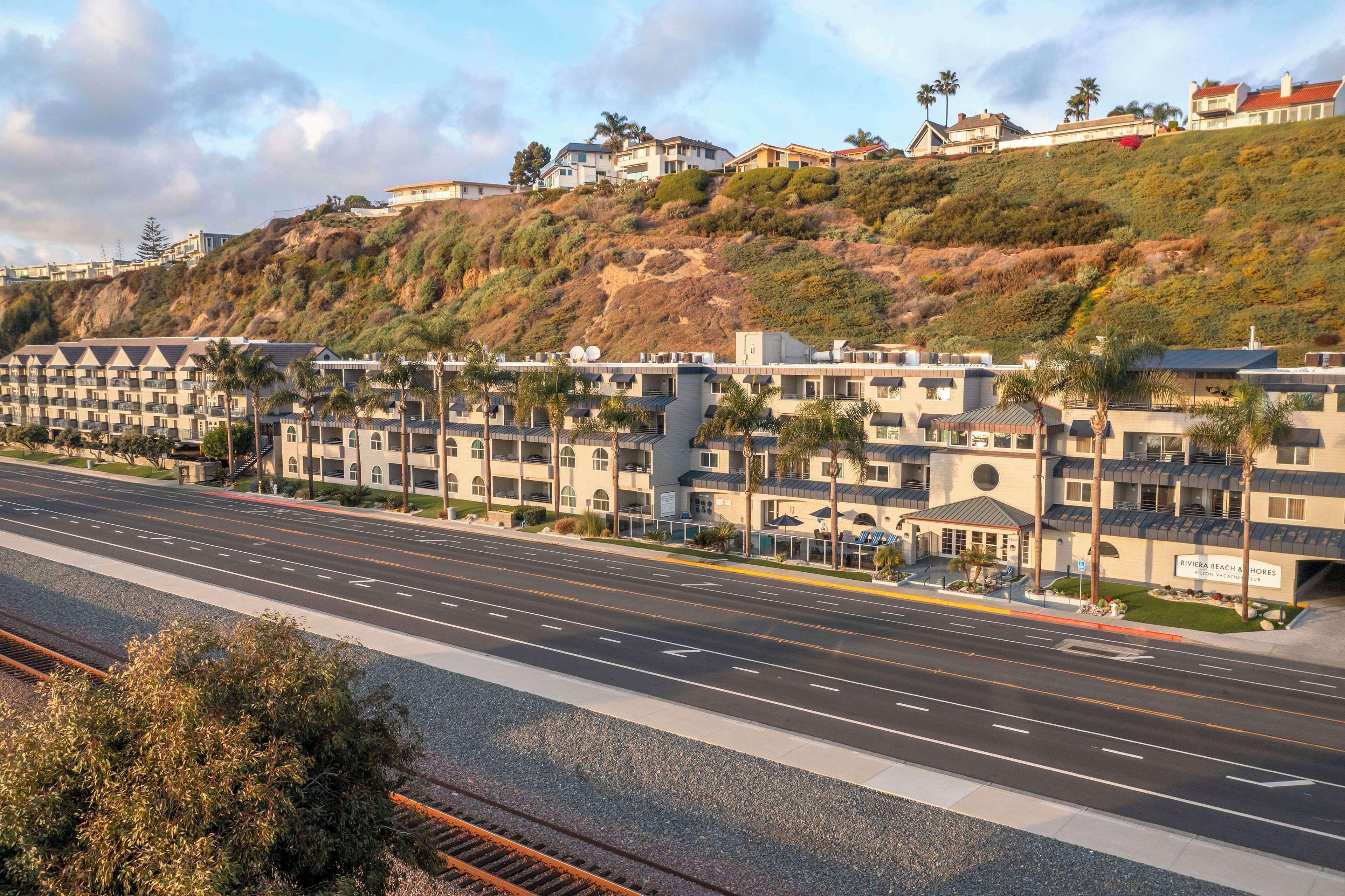A coastal hotel with multiple buildings, a road in the foreground, and a hillside with vegetation.