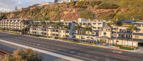 A coastal hotel with multiple buildings, a road in the foreground, and a hillside with vegetation.