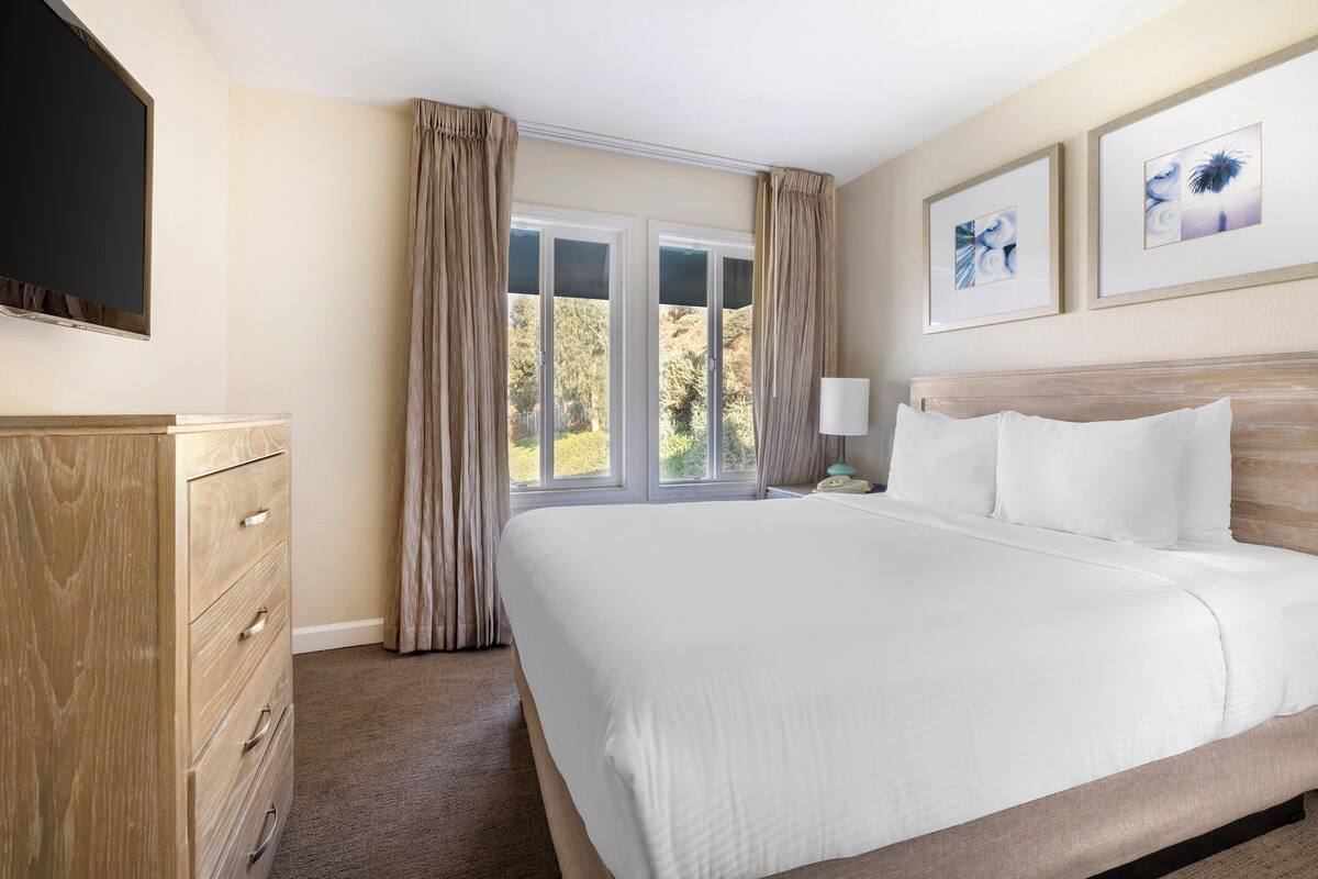 Ein Hotelzimmer mit einem großen Bett, einem Holzkasten, einem Flachbildfernseher und einem Fenster mit Vorhängen.