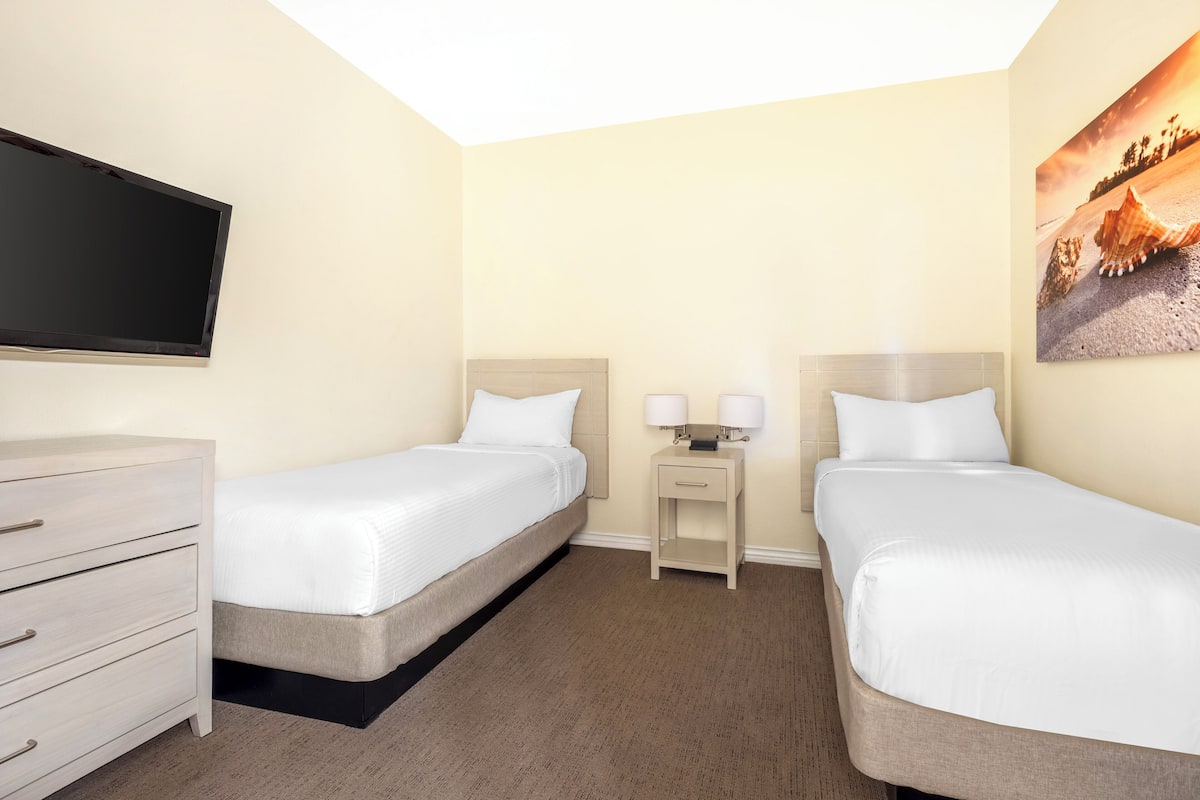 Ein Hotelzimmer mit zwei Betten, einem Fernseher, einem Schminktisch und einem Gemälde an der Wand.
