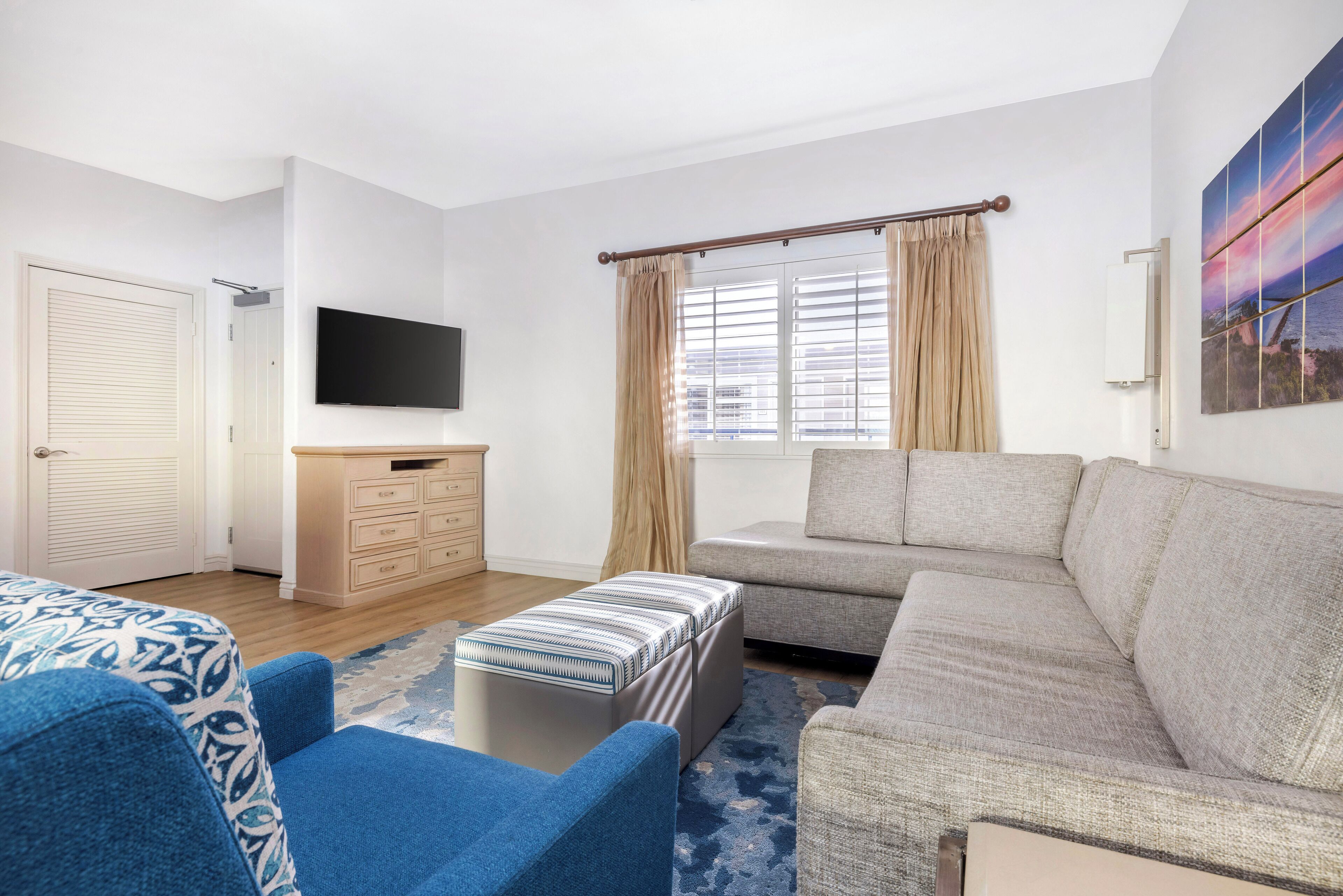 A modern hotel room with a sofa, a blue armchair, a wooden dresser, a flat-screen TV, and a large window with curtains.