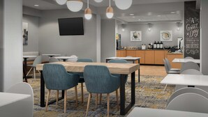 A modern dining area with a long wooden table, blue upholstered chairs, and pendant lighting. - TownePlace Suites by Marriott Suffolk Chesapeake (Suffolk)