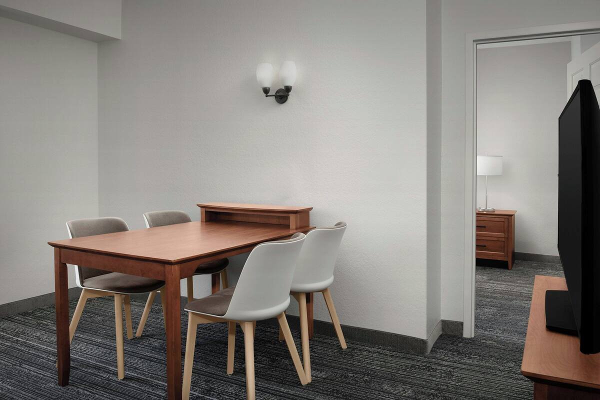 A conference room with a wooden table, four chairs, a wall-mounted light fixture, and a television on a stand.
