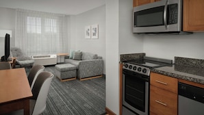 A modern kitchen with a microwave, stove, and dishwasher. There is a dining area with a table and chairs, and a living area with a sofa and a coffee table. - TownePlace Suites by Marriott Suffolk Chesapeake (Suffolk)