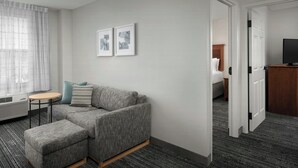 A modern hotel room with a grey sofa, a small round table, and a flat-screen TV. - TownePlace Suites by Marriott Suffolk Chesapeake (Suffolk)