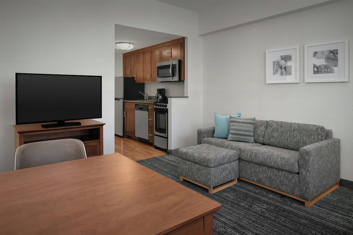 A modern living room with a wooden dining table, grey sectional sofa, and a flat-screen TV.