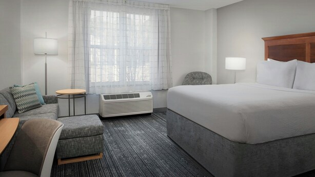 A hotel room with a bed, a sofa, a chair, a small table, and an air conditioning unit. - TownePlace Suites by Marriott Suffolk Chesapeake (Suffolk)