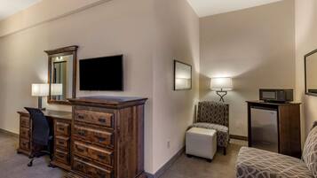 Una habitación de hotel con un aparador de madera, un televisor de pantalla plana, un escritorio con una silla, un sillón estampado, un pequeño sofá y un refrigerador.