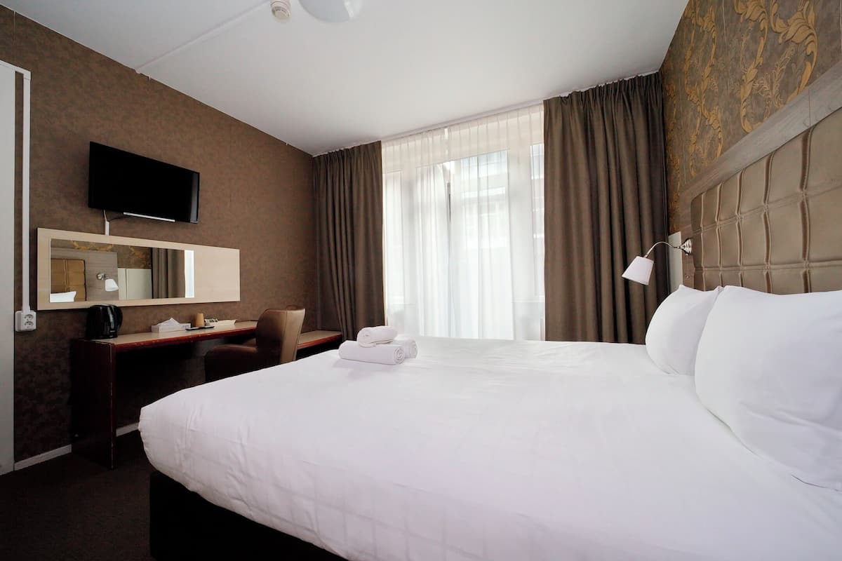 Une chambre d’hôtel avec un grand lit, un bureau avec une chaise, un téléviseur à écran plat fixé au mur et une fenêtre avec des rideaux.