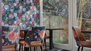 Een eetgedeelte met stoelen en tafels met bloemenmotief, een houten hekwerk zichtbaar door het raam, en een uitzicht op bomen.
