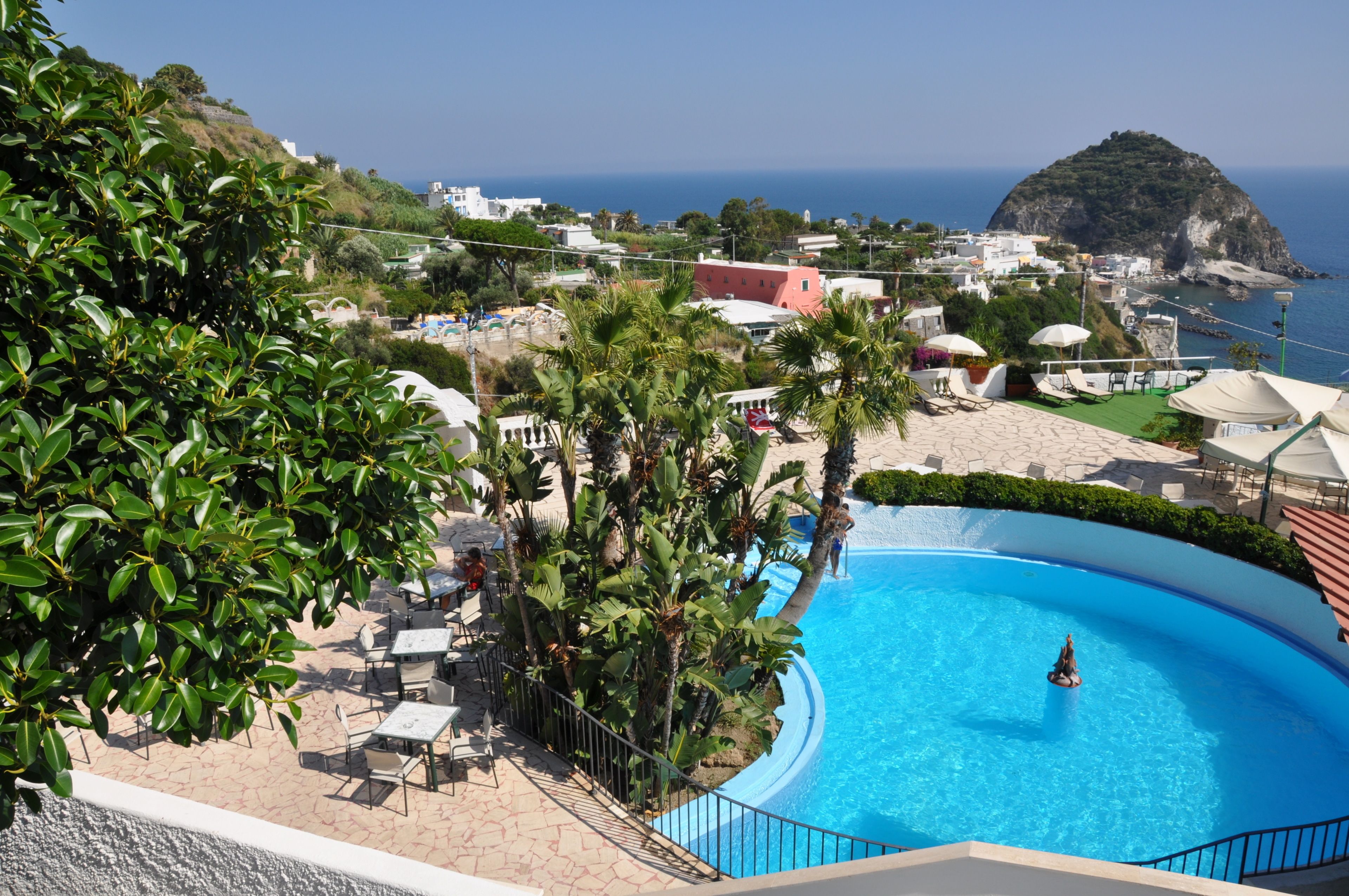 A resort with a swimming pool, outdoor seating, and a view of the sea and a hill.