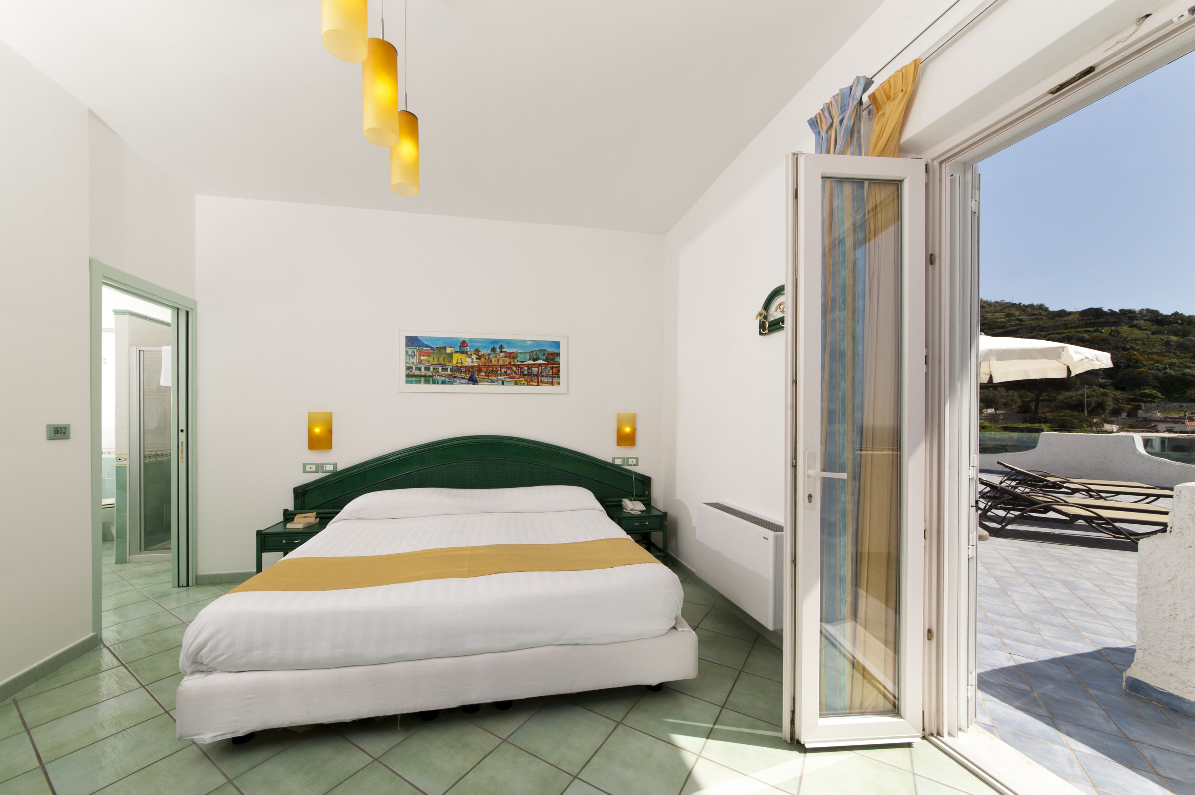 Habitación de hotel con cama, un cuadro, mesita de noche, teléfono y puerta corrediza de vidrio que da a un área exterior con sombrillas.