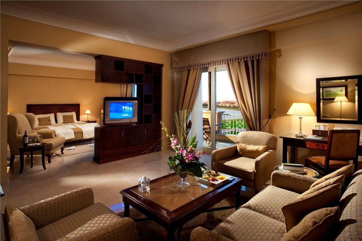 Une chambre d’hôtel avec un lit, une télévision, une table basse, un canapé et un fauteuil. Il y a une fenêtre avec des rideaux et une vue sur le paysage urbain.