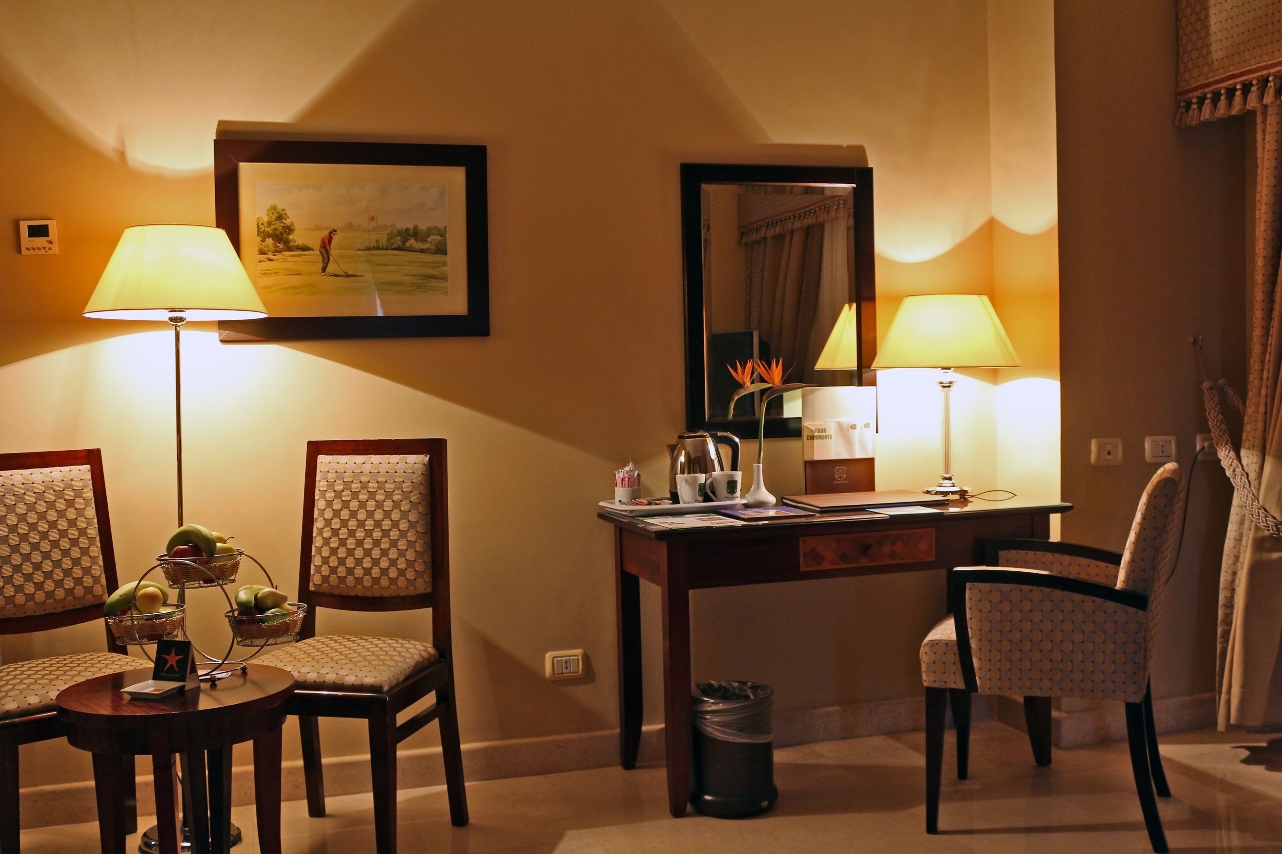 Ein Hotelzimmer mit Schreibtisch, zwei Stühlen, einer Lampe, einem Mülleimer und einem gerahmten Bild an der Wand.