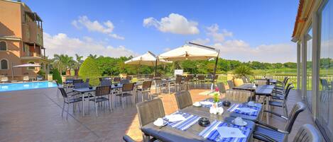 An outdoor dining area with tables and chairs, a pool, and a view of greenery and buildings.