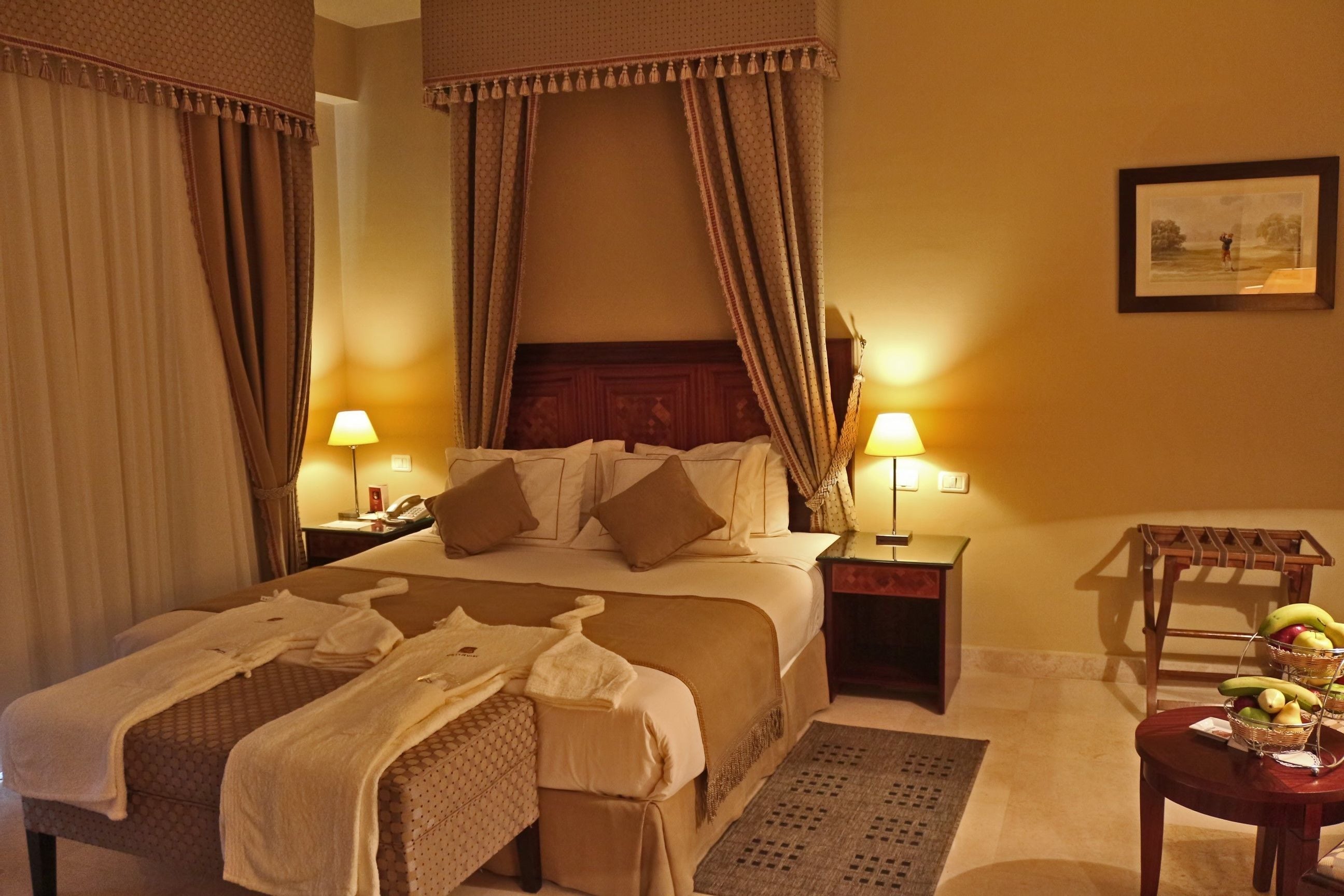 A hotel room with a large bed, two bedside tables with lamps, a wooden side table with fruit, and a framed picture on the wall.