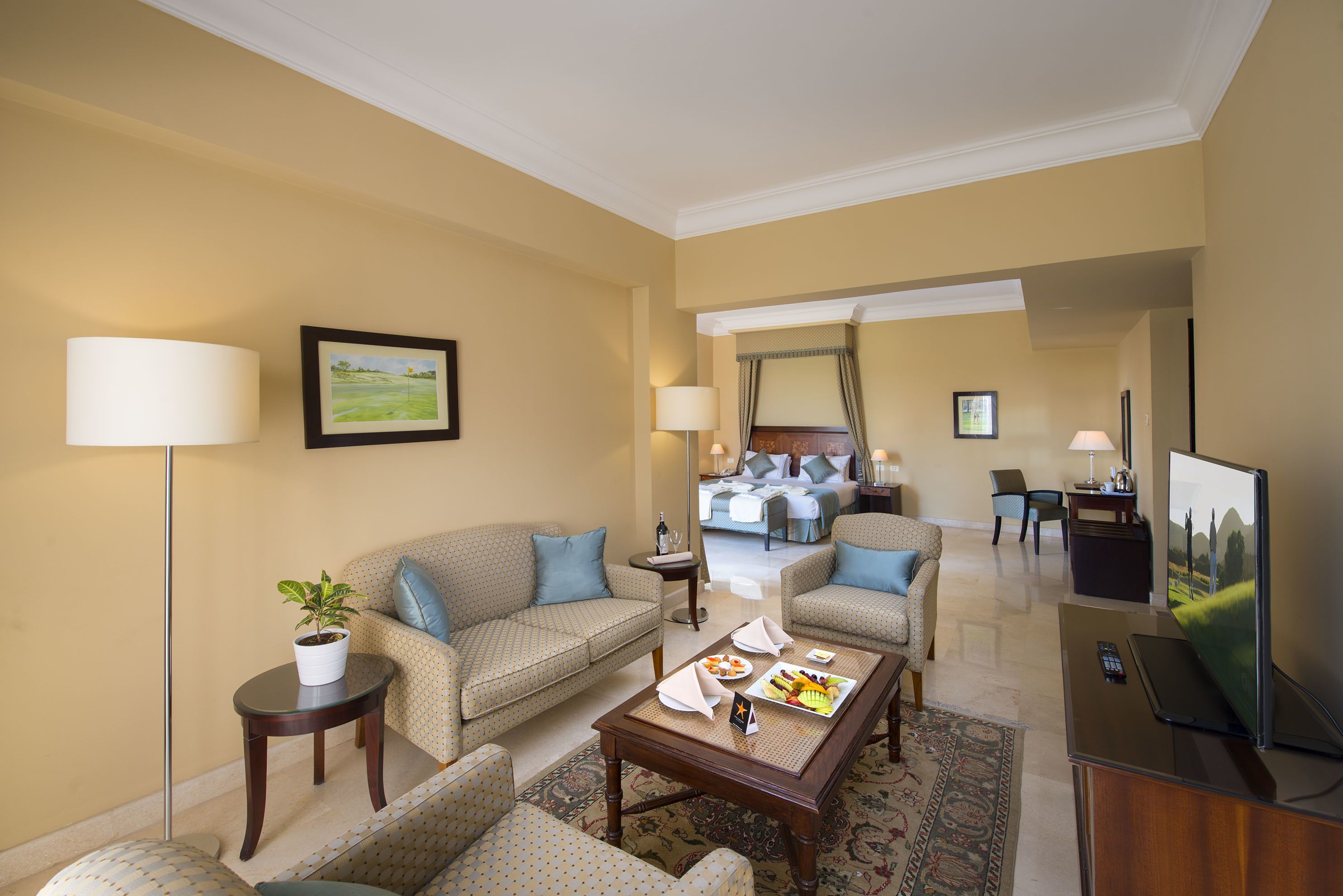 A hotel room with a living area featuring a sofa, armchairs, a coffee table, and a TV. There is a bed in the background with a view of the outdoors.