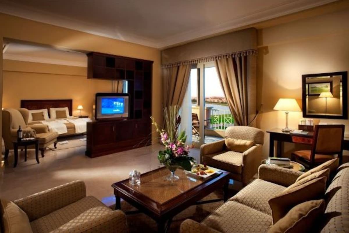 Quarto de hotel com uma cama grande, duas poltronas, um sofá, uma TV, uma mesa de centro e vista para o exterior.
