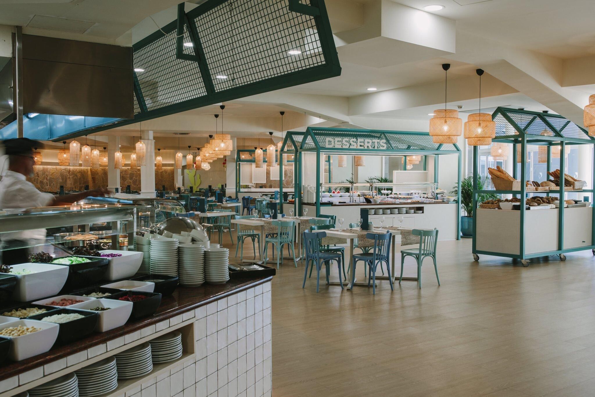 A modern restaurant with a central dessert station, multiple dining areas with blue chairs, and a chef preparing food.