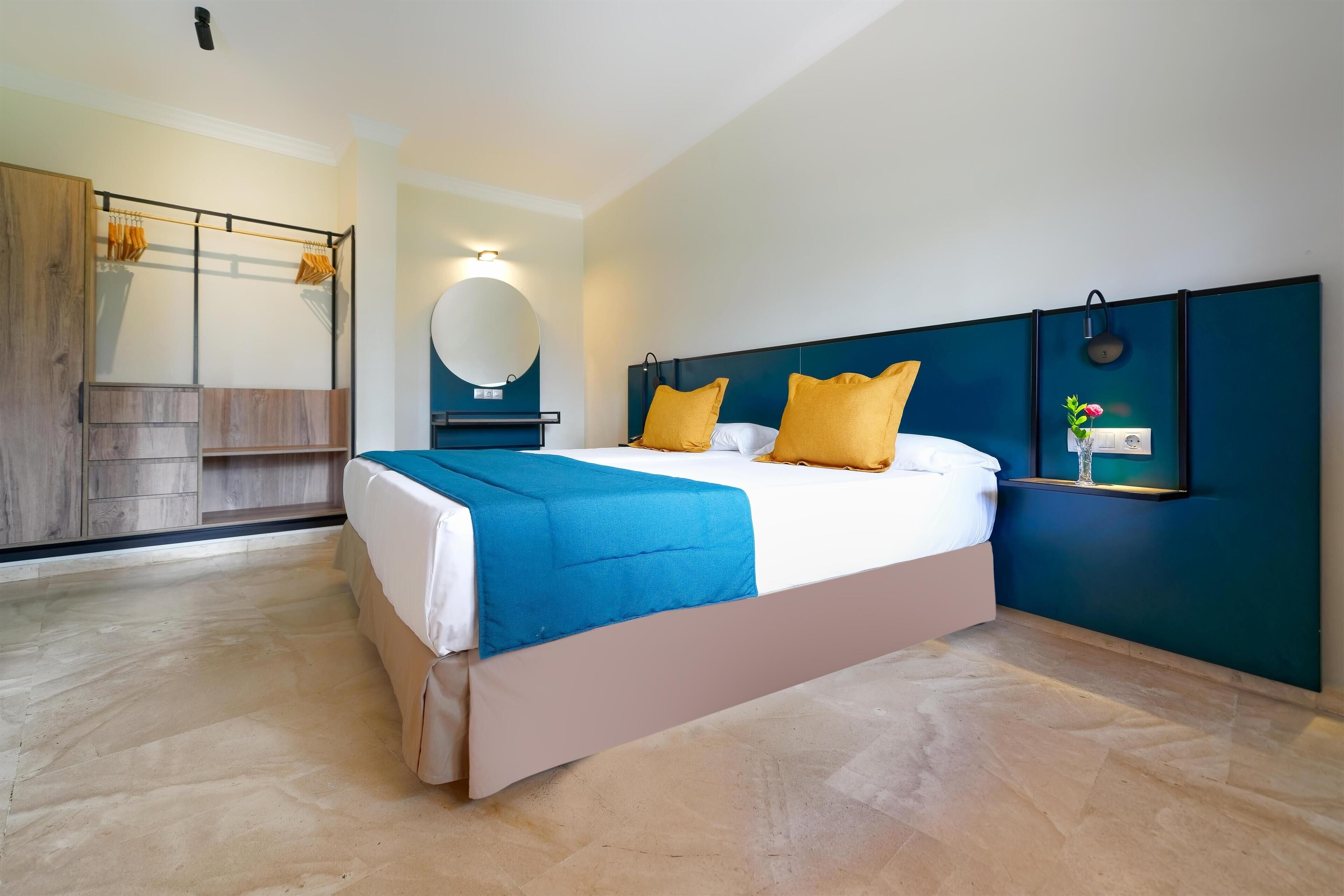 Una habitación de hotel moderna con una cama grande, un armario de madera, un espejo redondo y un estante pequeño con objetos decorativos.