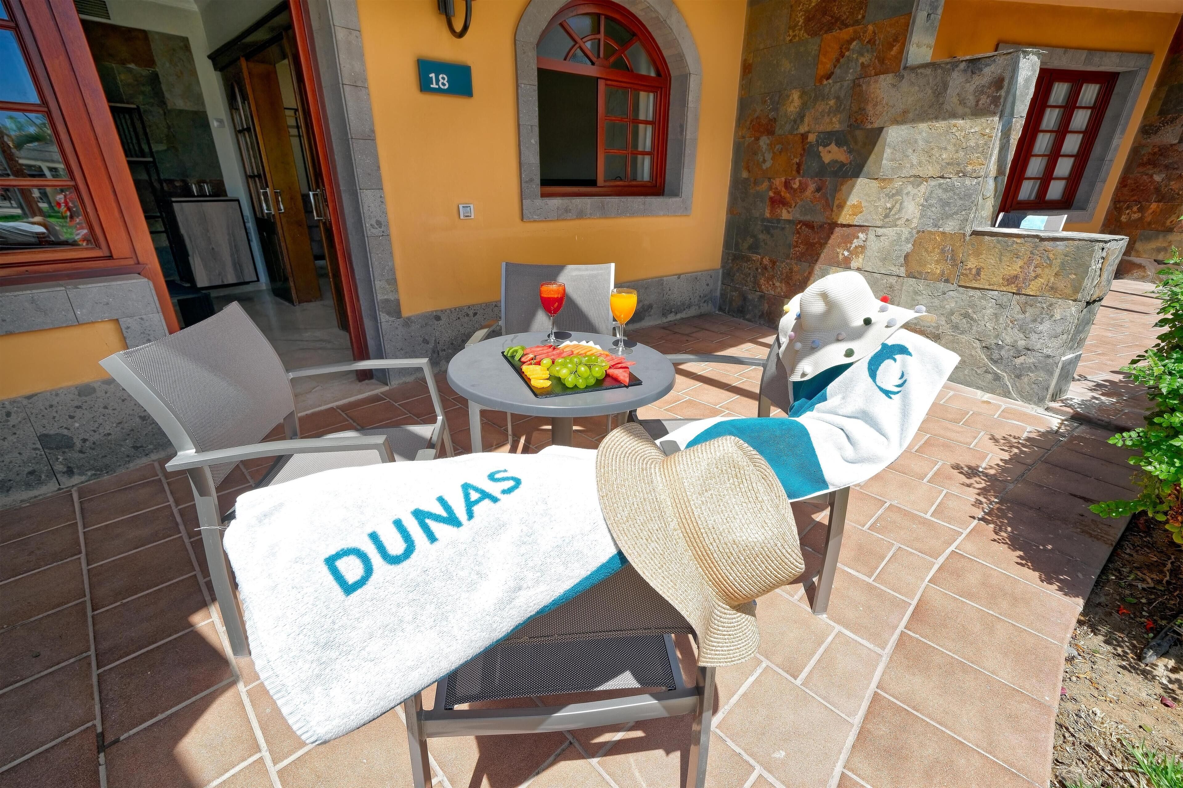 A sunlit patio with a table set for two, a chair with a towel labeled "DUNAS," and a fruit platter with drinks.