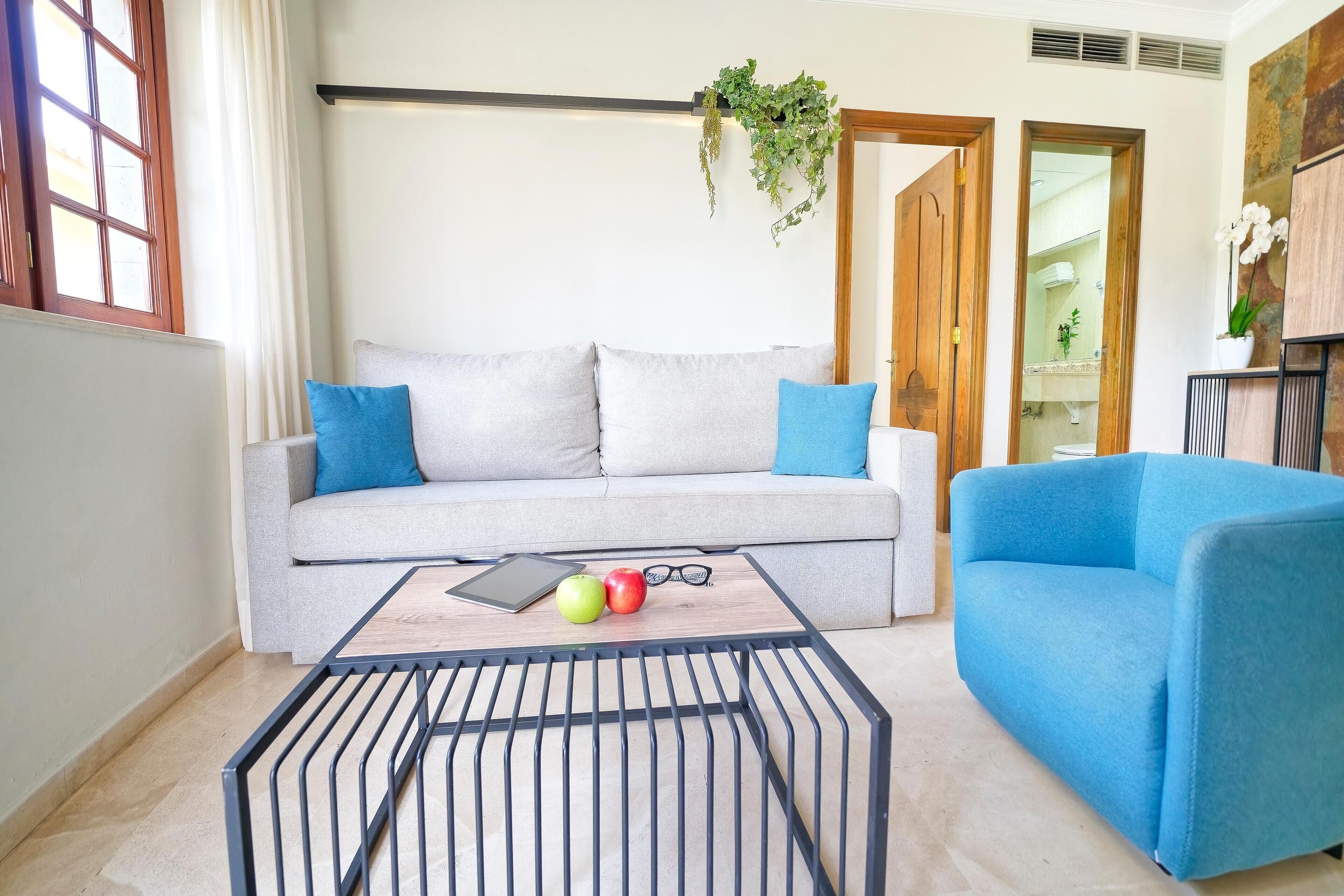 Ein Wohnzimmer mit grauem Sofa, blauen Kissen, einem blauen Sessel, einem Couchtisch mit einem Tablet, Äpfeln und Gläsern.