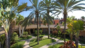A courtyard with palm trees, a pathway, and a building with a tiled roof. - Suites & Villas by Dunas (San Bartolomé de Tirajana)
