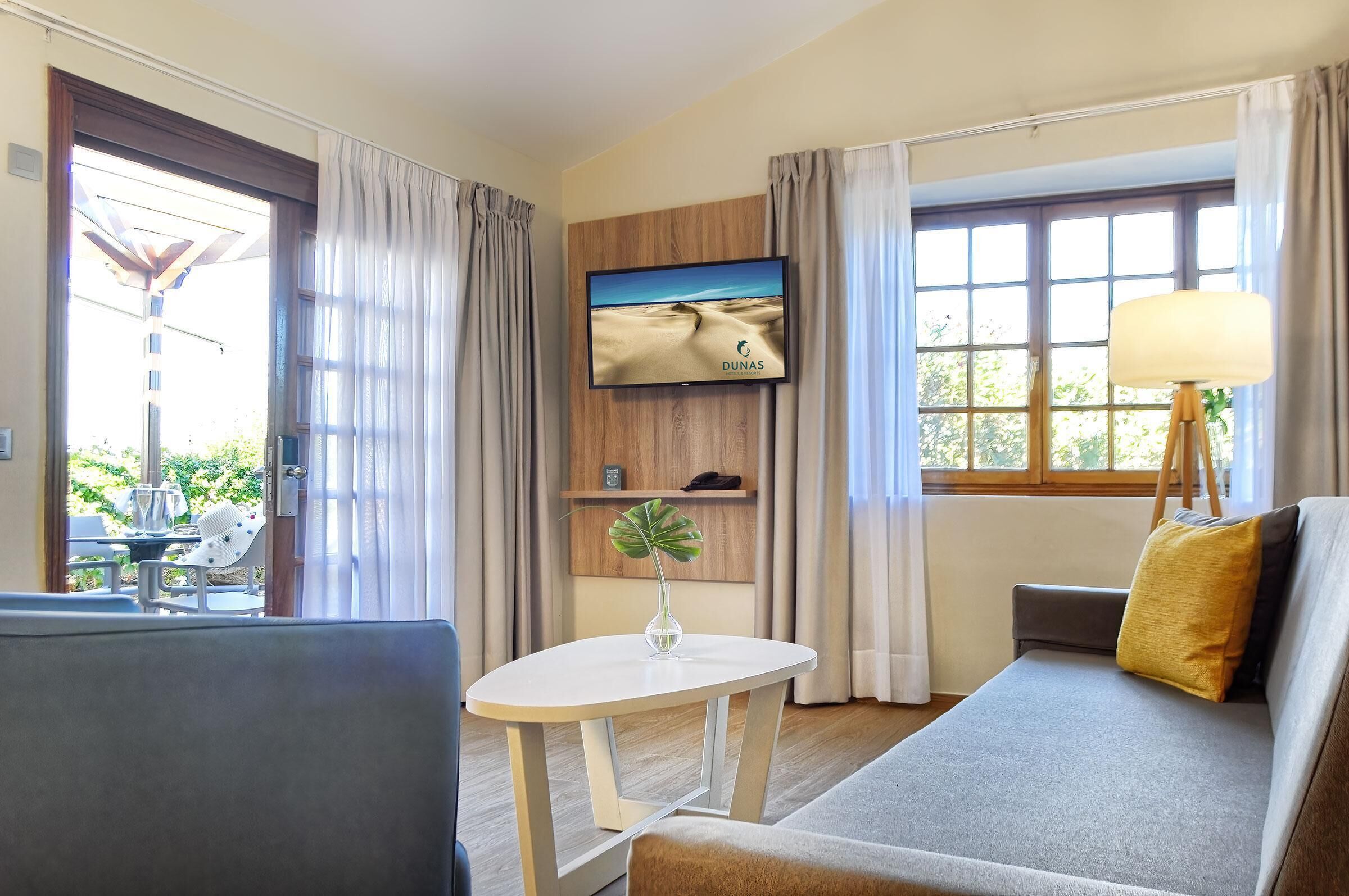 Habitación de hotel con sofá, una mesita con un jarrón, televisión en la pared y ventana con cortinas.