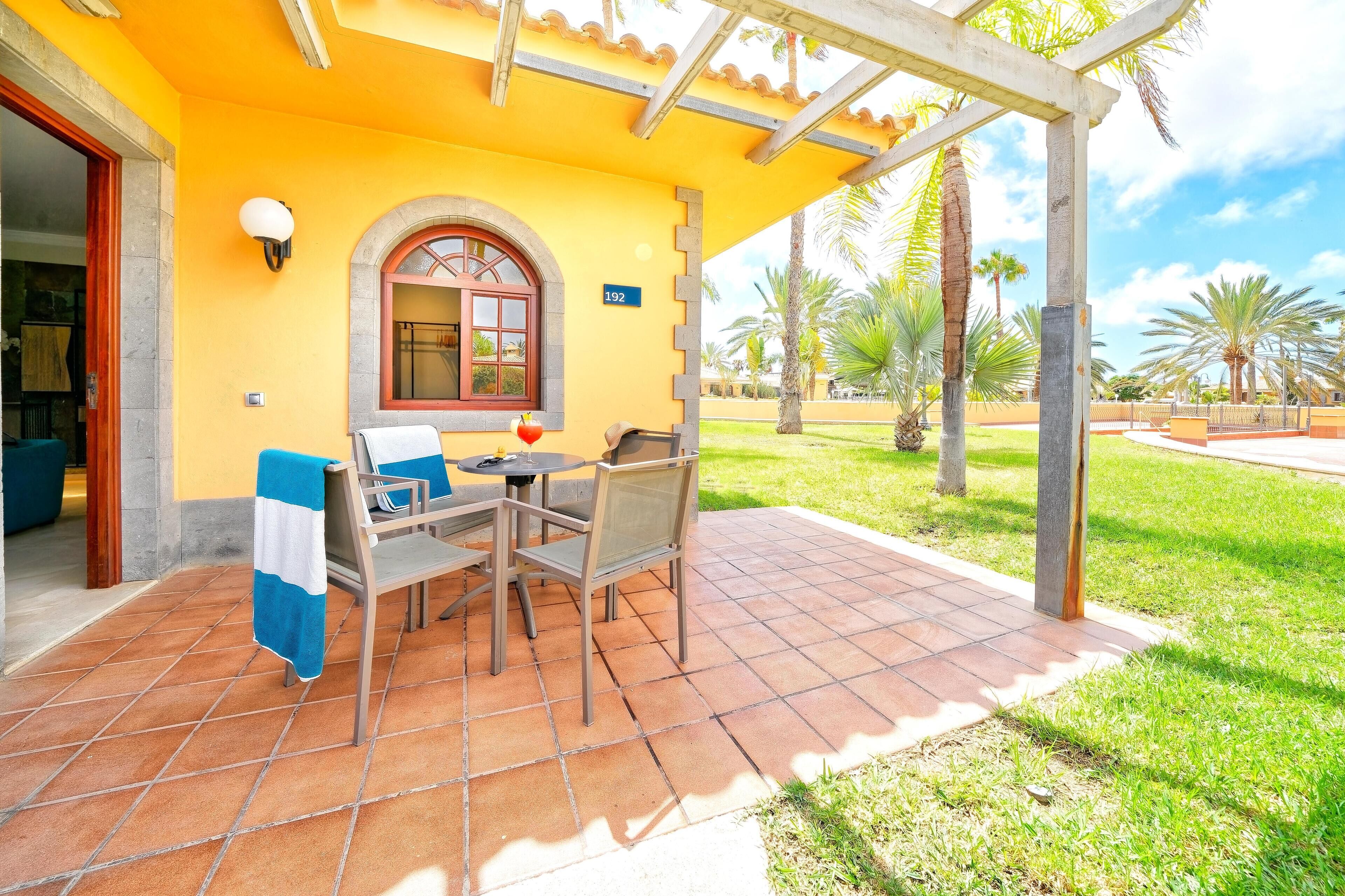 A patio with a table and chairs, a covered seating area, and a view of a well-maintained lawn and palm trees.