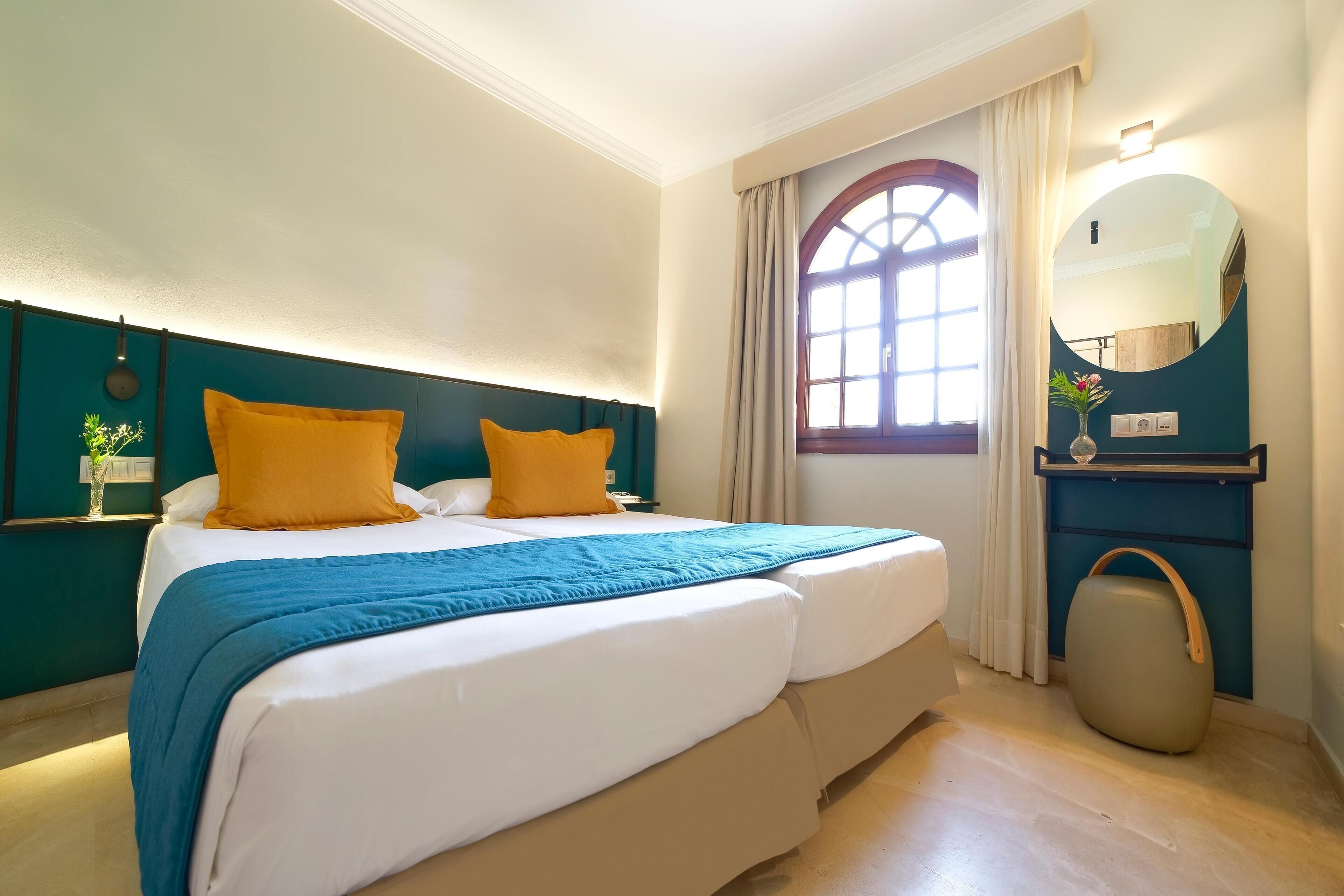 A hotel room with a large bed, two yellow pillows, a blue blanket, a wooden headboard, a round mirror, a small plant, and a window with curtains.