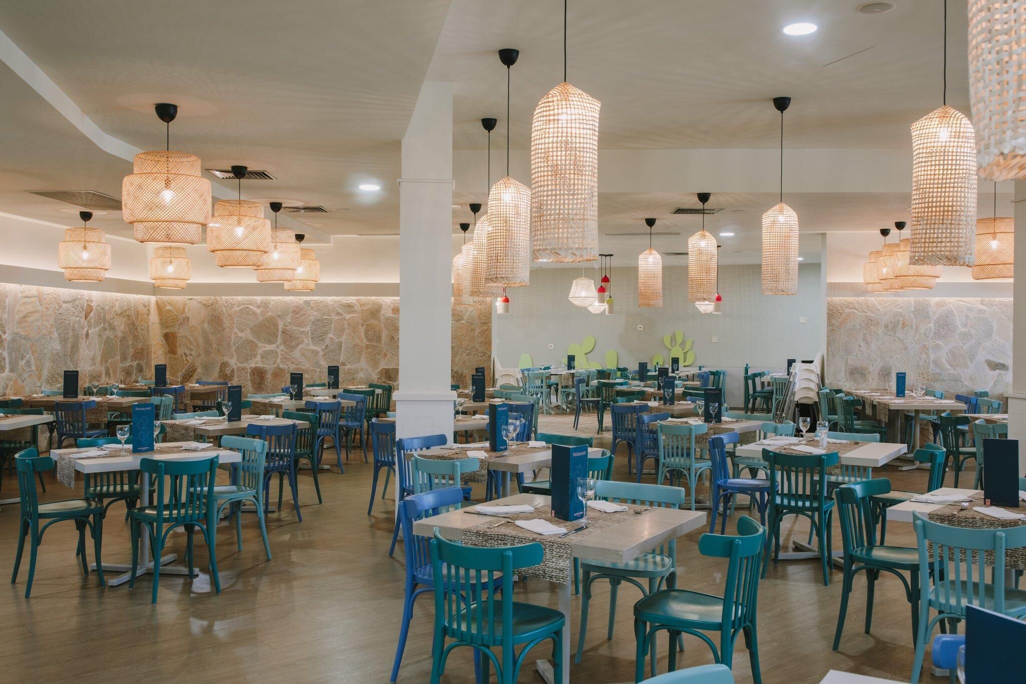A spacious restaurant with multiple tables and chairs, featuring a stone wall and hanging wicker light fixtures.